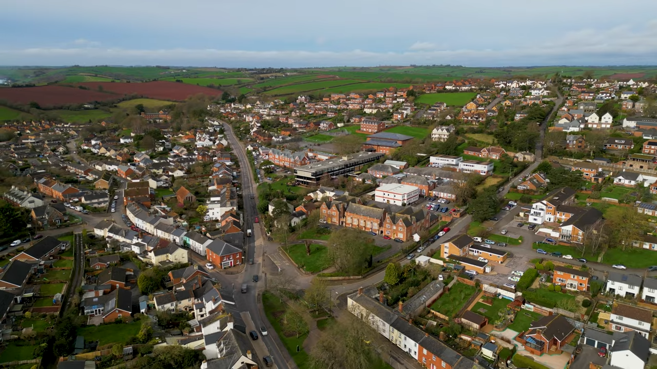 Aerial view of Queen Elizabeth's School, Crediton, Western Road campus with a winding road, surrounded by clusters of houses. Green fields and hills stretch into the distance under a cloudy sky.