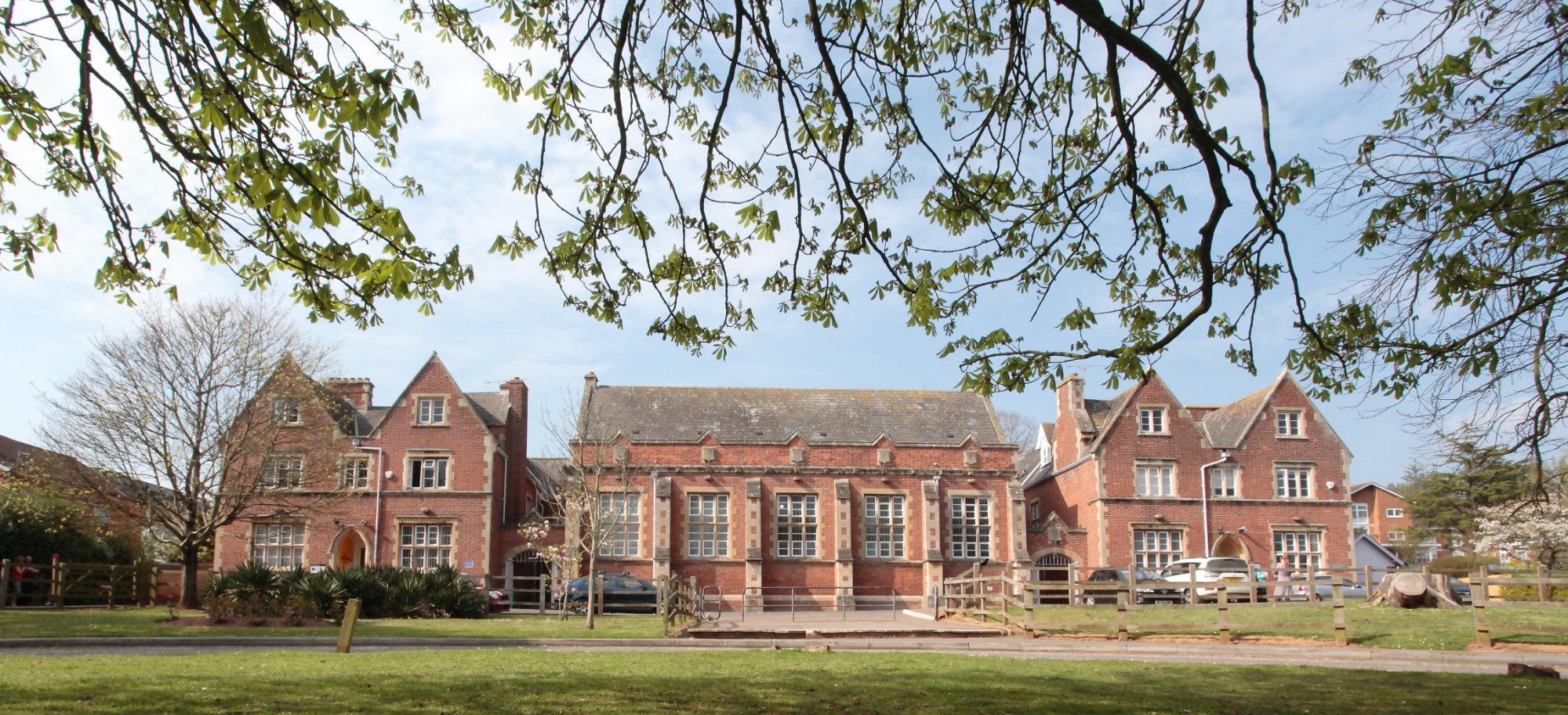 College House, Queen Elizabeth's School Western Road Campus is a large red-brick building with arched windows and peaked roofs, it sits in a lush green yard. Overhanging tree branches frame the sunny, peaceful scene.